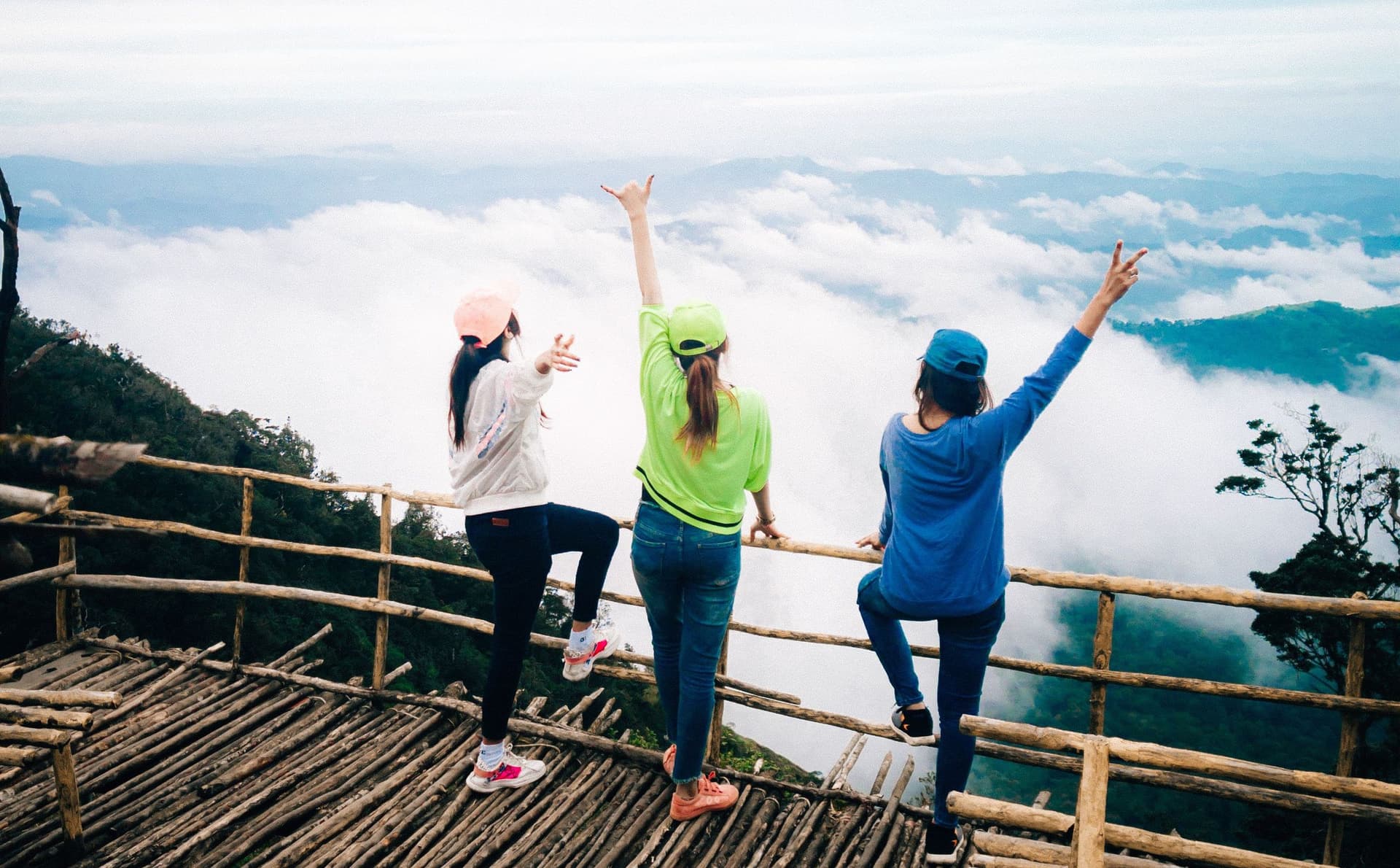 Three young women with raised arms on a hilltop viewpoint above the clouds in Kerala